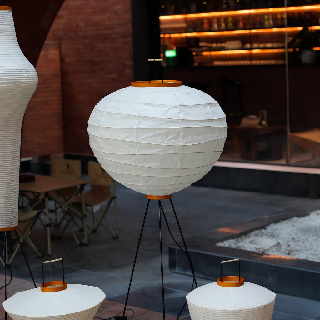 Akari 10D Floor Lamp — shade on a tripod on a restaurant terrace beside cafe tables and gravel — Captains Bamboo