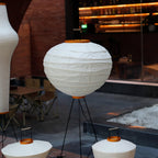 Akari 10D Floor Lamp — shade on a tripod on a restaurant terrace beside cafe tables and gravel — Captains Bamboo