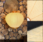Akari 3A Table Lamp — lit lamp against a wall of stacked firewood, paper rib insets — Captains Bamboo