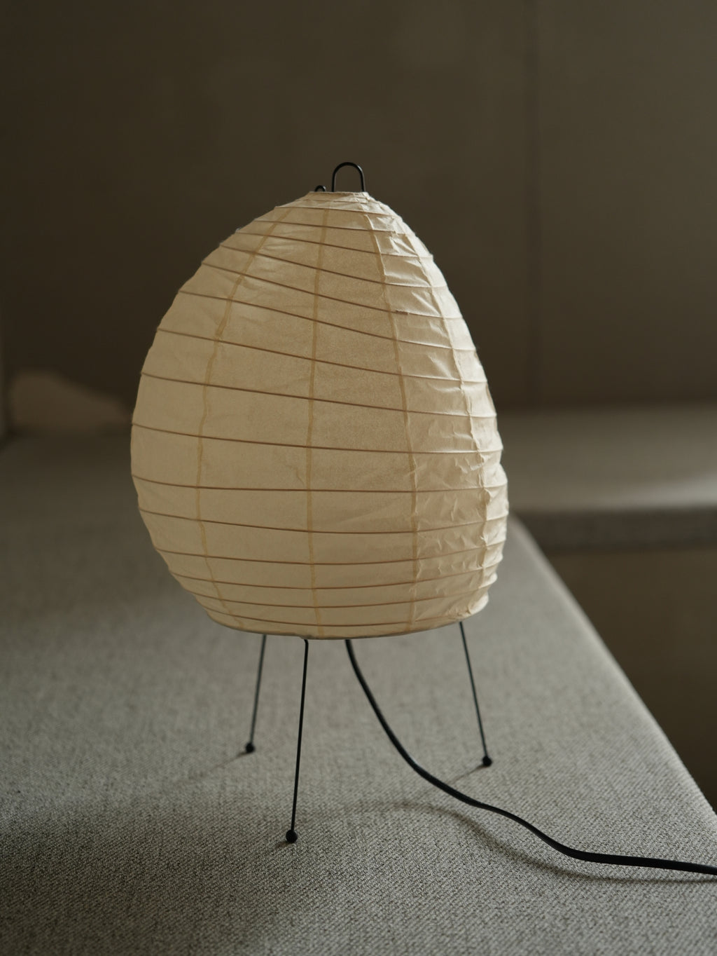 Akari 1N Table Lamp — unlit shade on a linen bench in soft window light, tripod legs in view — Captains Bamboo