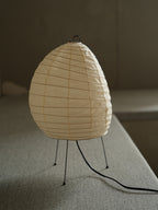 Akari 1N Table Lamp — unlit shade on a linen bench in soft window light, tripod legs in view — Captains Bamboo