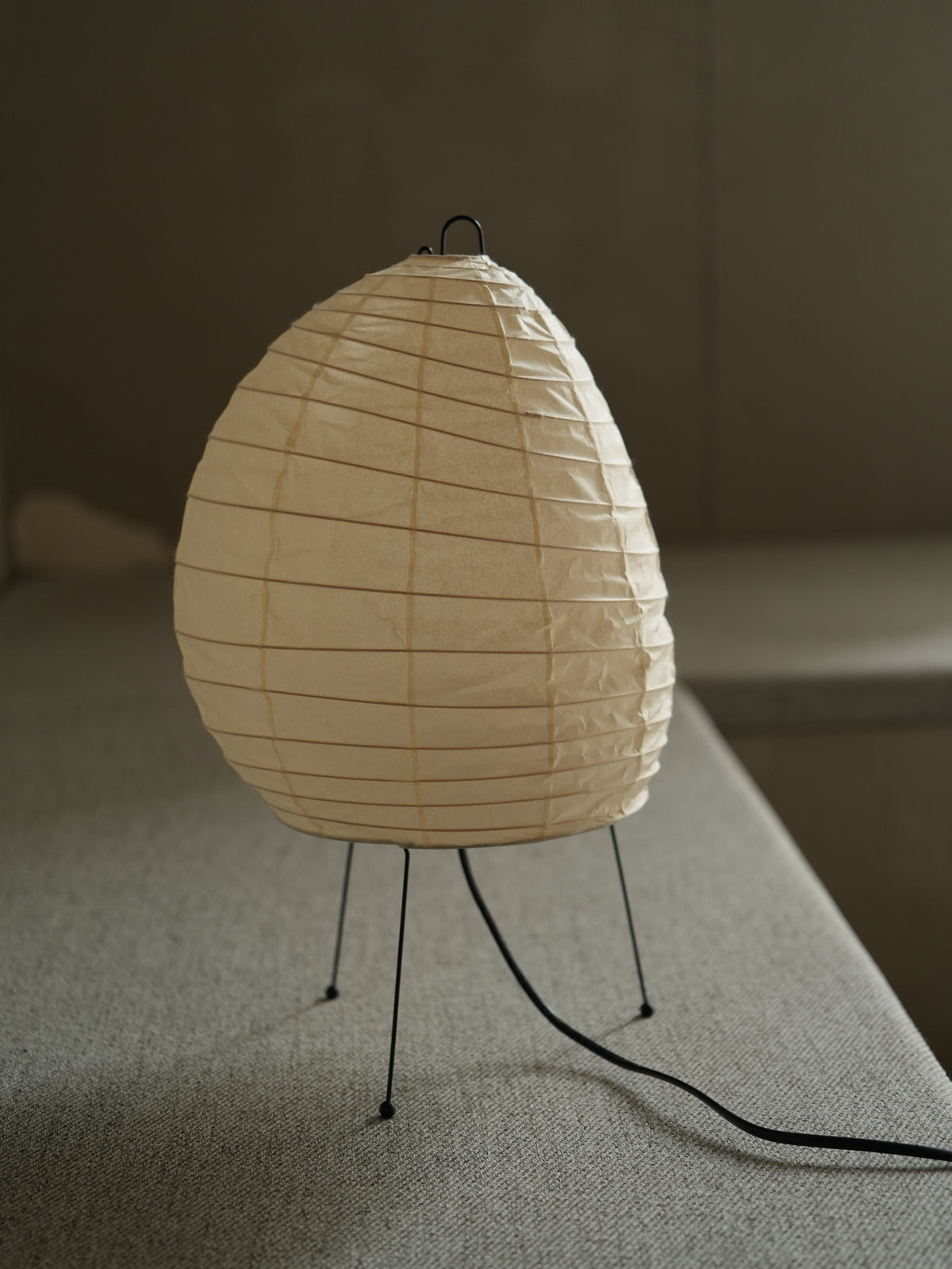 Akari 1N Table Lamp — unlit shade on a linen bench in soft window light, tripod legs in view — Captains Bamboo
